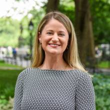 Jen is outside in a park with trees behind her. She is wearing a black and white blouse.